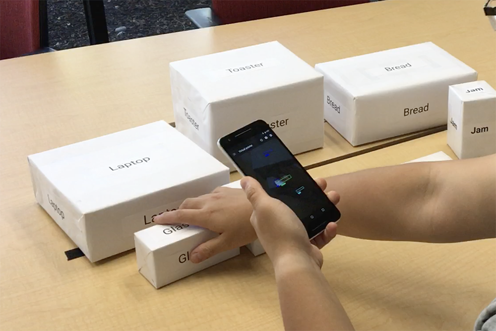 A table with many boxes covered with white paper showing text such as glasses, butter, jam, etc. A user is holding and targeting his phone at one object, while touching the object with the other hand. This is showcasing the window cursor interaction technique that supports non-visual attention to items within a complex visual scene, in which the user moves the device itself to scan the scene and receives information about what is in the center of the image.