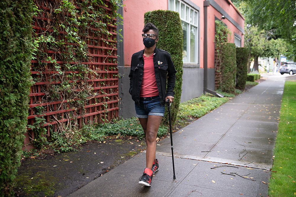 Leila, a black, non-binary person with a filtering face mask walks down a neighborhood street with one hand in their pocket and the other hand on their cane. They have a short mohawk and are wearing a jacket, shorts, tennis shoes, and glasses.