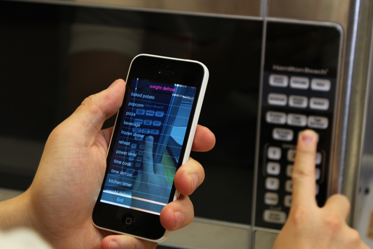 The user is holding the phone in portrait mode with one hand, and aiming the camera towards an inaccessible microwave control panel. The user’s other hand is exploring on the panel. The VizLens iOS app is providing audio feedback and guidance to the user.